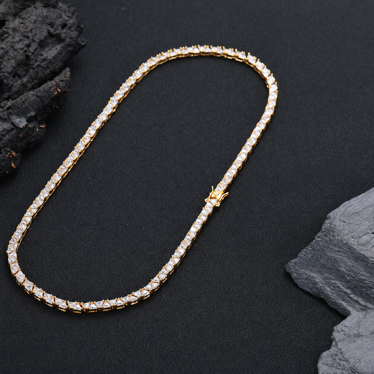 Gold-tone triangle tennis necklace laid in a long loop beside dark rocks