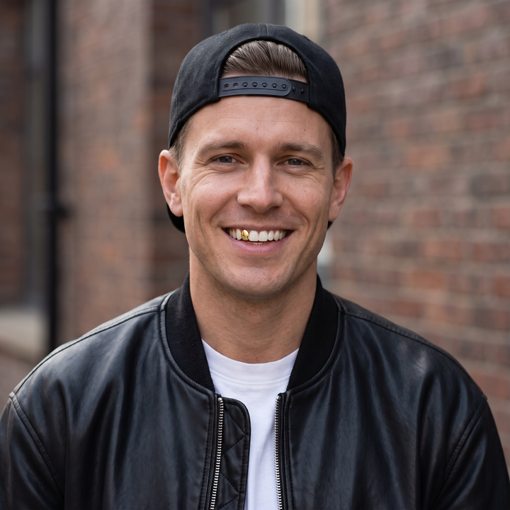 Smiling model in a backward black cap and leather jacket wearing one gold-tone tooth grill