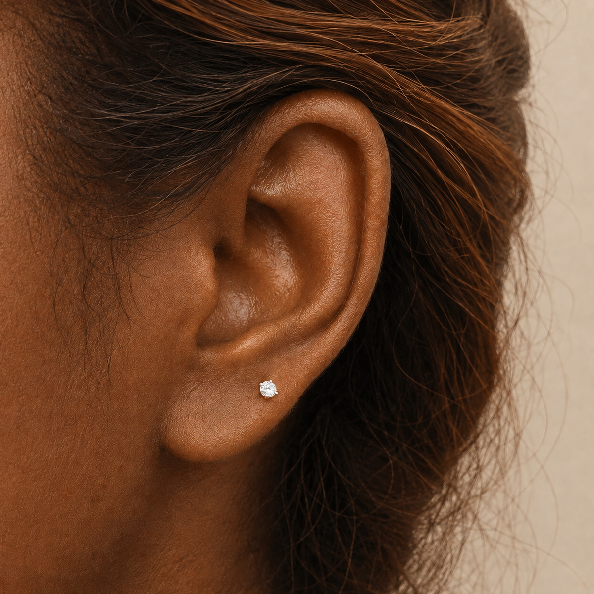 A close-up of a person's ear wearing a small 3mm round stud earring, with dark brown hair and a soft beige background.
