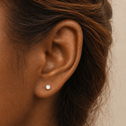 A close-up of a person's ear wearing a small 4mm round stud earring, with dark brown hair and a soft beige background.